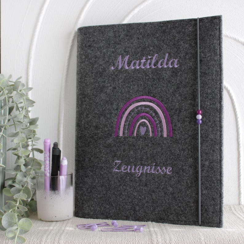 Op de afbeelding: Een donkergrijze vilten map met de geborduurde naam Matilda, een paarse regenboogafbeelding en het woord Zeugnisse. De map wordt gesloten met een verticaal elastiek met drie kleine paarse kralen en staat naast een glazen pennenhouder.