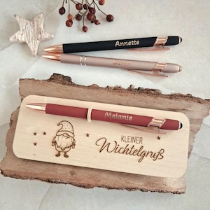 Op de afbeelding: Drie balpennen met roségouden accenten. Eén zwarte pen gegraveerd met Annette, één roségouden pen gegraveerd met Sarah en één matte rode pen gegraveerd met Melanie op een houten plaat met een kabouter en de tekst Kleiner Wichtelgruß.