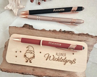 May include: Three ballpoint pens with rose gold accents. One black pen engraved with Annette, one rose gold pen engraved with Sarah, and one matte red pen engraved with Melanie resting on a wooden plaque with a gnome and Kleiner Wichtelgruß text.