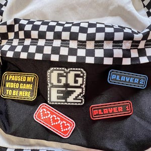 Op de afbeelding: Zwart-wit geruite rugzak met patches. Patches zijn onder meer "I paused my video game to be here", "GG EZ", "Player 1" en "Player 2". De rugzak heeft ook een "Off The Wall" patch.