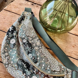 Könnte beinhalten: Eine gemusterte Bauchtasche mit grünem Riemen und Reißverschluss. Die Tasche hat ein Blumenmuster in Grün-, Weiß- und Brauntönen. Der Reißverschluss ist goldfarben. Im Hintergrund befindet sich eine grüne Glasvase.