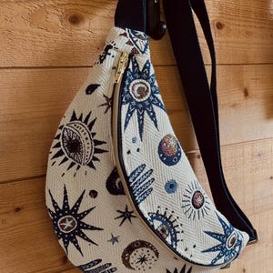 Könnte beinhalten: Eine weiße Bauchtasche mit einem schwarzen verstellbaren Gurt. Die Bauchtasche hat einen goldenen Reißverschluss und ein buntes Muster aus Sternen, Monden, Augen und Händen.