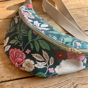 Könnte beinhalten: Eine dunkelgrüne Bauchtasche mit Blumenmuster. Die Tasche hat einen Reißverschluss und einen verstellbaren beigefarbenen Gurt.
