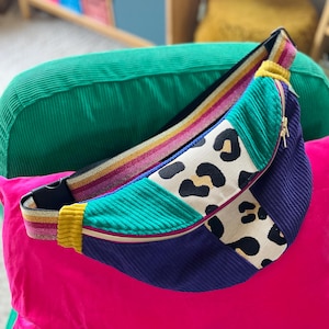 Puede incluir: Una riñonera multicolor con un panel de estampado de leopardo, un panel de pana verde, un panel de pana azul y una correa a rayas amarilla y rosa.