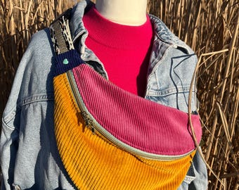 May include: A mustard yellow, pink, and blue corduroy fanny pack with a zipper closure. The fanny pack is worn on a denim jacket.