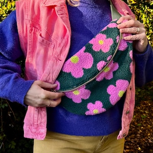 May include: A pink denim vest over a blue sweater. A green and pink floral print fanny pack with a silver zipper is being held. The fanny pack has a repeating pattern of pink flowers with yellow centers.