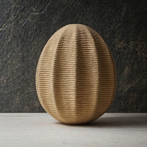 Peut inclure: Un objet décoratif beige en forme d'œuf avec une surface texturée et nervurée. L'objet est posé sur une surface de couleur claire avec un fond gris foncé.