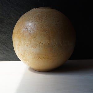 Op de afbeelding: Een grote, bolvormige, beige keramische vaas met een textuur. De vaas heeft een afgeronde vorm en een kleine opening aan de bovenkant. De achtergrond is donker, waardoor de vorm van de vaas wordt benadrukt. De vaas staat op een wit oppervlak.