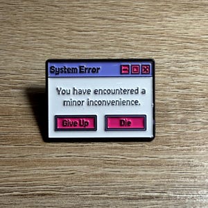 May include: A rectangular enamel pin featuring a purple and white computer system error window. The text reads System Error, You have encountered a minor inconvenience, Give Up, and Die. The design includes pink buttons and a black border.