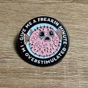 May include: Circular enamel pin depicting a pink pufferfish with crying eyes. The pin has a light blue background and a black border with the text "GIVE ME A FREAKIN MINUTE I'M OVERSTIMULATED".