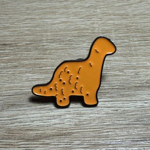 May include: An orange dinosaur enamel pin with a black outline. The pin features a stylized design of a long-necked dinosaur with small, dark spots on its body. The pin is set against a wooden background.