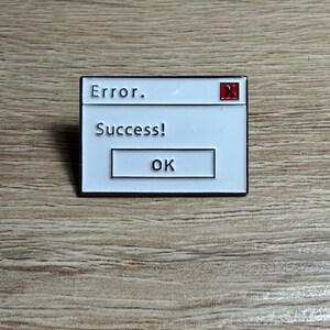 May include: A white enamel pin with a black border, styled as a computer error message. The text reads "Error." above "Success!" and "OK" in a button. A red "X" is in the top right corner.