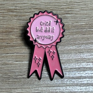 May include: A pink enamel pin in the shape of a ribbon with the text "Cried But did it anyway". The pin has a black outline and features sparkling star accents. The ribbon has two streamers.