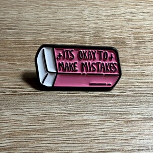 May include: A rectangular enamel pin shaped like a pink and white eraser with black outlines. The pin features the text IT'S OKAY TO MAKE MISTAKES in black lettering across the pink section, with small black plus signs on either side of the text.