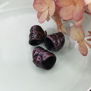 Könnte beinhalten: Drei kleine, dunkelviolette und schwarze Metallperlen mit einer strukturierten Oberfläche. Die Perlen haben die Form eines Zylinders mit abgerundeter Ober- und Unterseite.