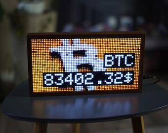 May include: A rectangular digital LED display screen in a dark wooden frame showing a white Bitcoin symbol, the text BTC, and the numerical value 83402.32 with a dollar sign, all rendered in bright white pixels against a glowing orange background.