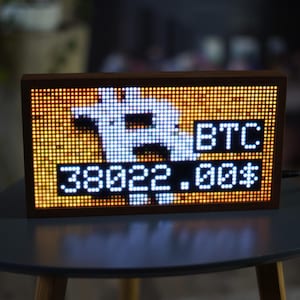 Peut inclure: Un écran LED rectangulaire avec un cadre en bois affiche le symbole Bitcoin, l'abréviation "BTC" et le prix "38022.00$". L'écran est posé sur une petite table ronde grise.