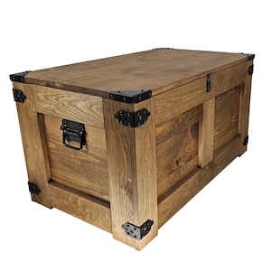 Op de afbeelding: Een houten opbergkist met een scharnierend deksel en zwarte metalen beslag. De kist heeft een rustieke bruine afwerking en is gemaakt van massief hout.
