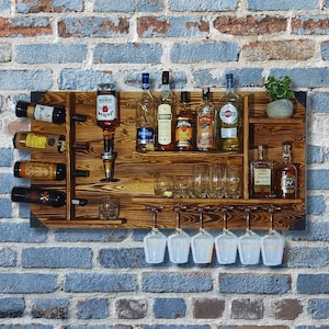 Può includere: Un'unità bar in legno a parete con ripiani per bottiglie e bicchieri. L'unità ha una finitura marrone rustica e presenta una varietà di bottiglie di liquore, tra cui bourbon, gin e vodka. Ci sono anche diversi bicchieri da vino appesi allo scaffale inferiore.