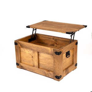 Puede incluir: Un cofre de almacenamiento de madera con tapa abatible. El cofre tiene un acabado de madera natural y herrajes de metal negro. La tapa está abierta y el interior del cofre es visible.
