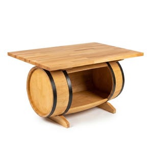 Peut inclure: Une table en bois brun clair dotée d'un plateau rectangulaire reposant sur une base en forme de tonneau horizontal. Le tonneau présente deux bandes métalliques noires et une section centrale ouverte formant une étagère. L'ensemble repose sur deux pieds.