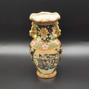 May include: A black and gold ceramic vase with a floral design and a scene of women in traditional Japanese clothing. The vase has two handles and a scalloped rim.