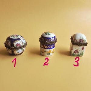 Op de afbeelding: Drie decoratieve porseleinen sieradendoosjes. De eerste is rond met een blauw en bloemenontwerp. De tweede is cilindrisch met blauwe en gele accenten. De derde is een kleine, witte doos met bloemendetails en een scharnierend deksel.