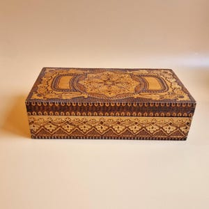 Può includere: Una scatola di legno con intagli intricati in marrone e oro. La scatola è rettangolare e ha un coperchio con cerniere. Gli intagli sono dettagliati e presentano un motivo floreale.