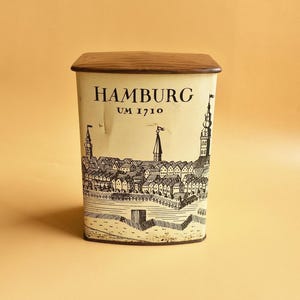 May include: A rectangular tin with a wooden lid. The tin is off-white with a black illustration of a city skyline and the word "HAMBURG" above "UM 1710". The lid is a dark brown color.
