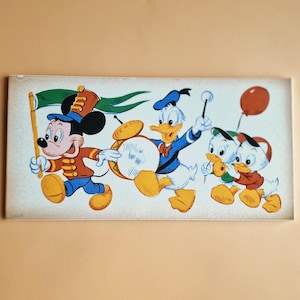 以下が含まれることがあります： ミッキーマウス、ドナルドダック、ヒューイ、デューイ、ルーイがパレードを行進している長方形のヴィンテージプリント。ミッキーマウスがパレードを先導し、ドナルドダックがドラムを演奏し、甥たちが風船を持っています。プリントは白い背景です。