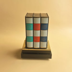 May include: A decorative desk ornament featuring three vertical book-shaped blocks on a black and gold base. The spines are white with colored panels in teal, red, and blue, each displaying Cyrillic text and dates including 1957 and 1966.