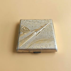 Puede incluir: Una pitillera cuadrada de metal dorado con un diseño espacial en relieve. La parte frontal muestra un cohete despegando hacia un cielo estrellado, con los textos 12-IV-1961, VOSTOK y CCCP grabados en la superficie de la caja metálica.