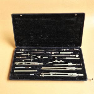Może przedstawiać: Zestaw do rysowania w stylu vintage w czarnym, wyściełanym aksamitem etui. Zestaw zawiera różne cyrkle, dzielniki i inne precyzyjne narzędzia do rysowania, wszystkie ze srebrnego metalu. Etui jest otwarte, ukazując uporządkowany układ instrumentów.