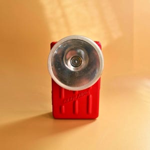 May include: A vintage red flashlight with a clear, circular lens and a silver reflector. The flashlight has a rectangular body with vertical lines and the word "Сделано" printed on the front. The background is a solid, light orange color.