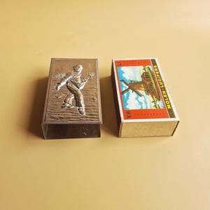 May include: A silver-colored match holder with a relief of a person walking in the snow next to a matchbox. The matchbox has a windmill illustration and the text "MOLEN-LUCIFERS".