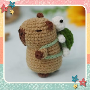 May include: A crocheted capybara carrying a turtle. The capybara is brown with dark brown accents and a green strap. The turtle is white with a green shell. The image includes the text "Capybara & Turtle" and "Cute Beginner Friendly Crochet Pattern".