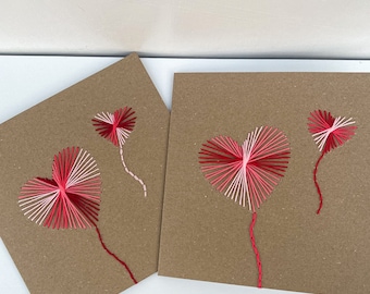 May include: Two handmade greeting cards with stitched heart designs. The hearts are crafted with red, pink, and white thread on a brown cardstock background. One card features two hearts, and the other has a single heart design.