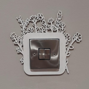 May include: White decorative light switch cover with a floral design. The cover is made of wood and has a cutout design of flowers and leaves. The switch cover is mounted on a light gray wall.