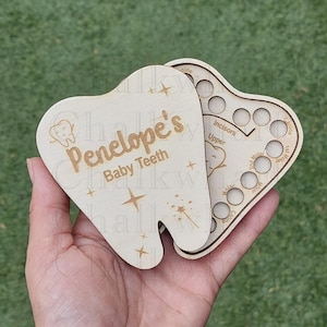 May include: A wooden tooth box shaped like a tooth with the name Penelope's Baby Teeth engraved on it. The box has a compartment for each tooth and is designed to hold baby teeth.