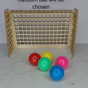 May include: A wooden game with a grid-like structure, resembling a goal, and five colourful balls: red, green, yellow, pink, and blue. The text "Random ball will be chosen" is above the game.