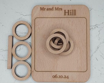 Könnte beinhalten: Holzsäulenspiel mit dem Text "Mr and Mrs Hill" und dem Datum "06.10.24" eingraviert. Das Spiel besteht aus einer Holzbasis mit einer quadratischen Öffnung und vier ineinandergreifenden Ringen.