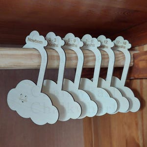 May include: Wooden hangers shaped like clouds with a smiley face. The hangers are labelled with age ranges in months: Newborn, 0-3, 3-6, 6-9, 9-12, 12-18.