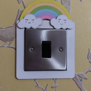 May include: A white light switch cover with a rainbow and two clouds with smiley faces. The rainbow is yellow, orange, red, purple, blue, and green. The clouds are white with black eyes and black smiles.