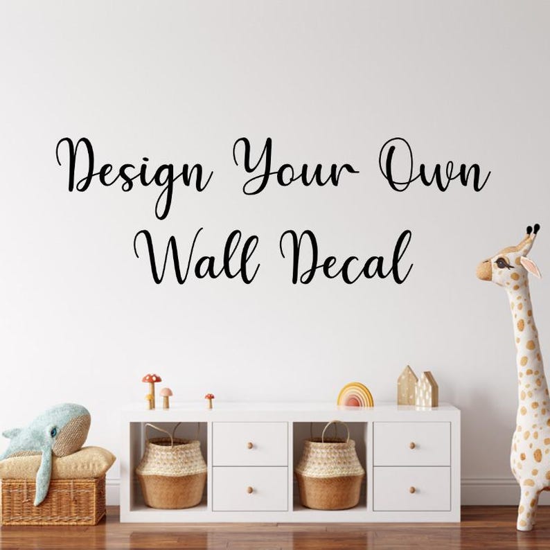 May include: A white wall featuring the text "Design Your Own Wall Decal" in black script. A white cabinet with woven baskets, a whale plush, and a giraffe decorates the room. A rainbow and wooden houses are also on the cabinet.