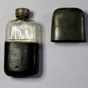 Könnte beinhalten: Eine Vintage-Flasche mit einem transparenten Glasaufsatz und einem schwarzen, lederummantelten Boden. Die Flasche hat einen Metallschraubverschluss und eine separate Metallabdeckung. Die Flasche ist rechteckig mit abgerundeten Kanten.