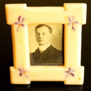 May include: A small, rectangular, cream-colored picture frame with a black and white photograph of a man. The frame is decorated with purple and yellow floral accents. The frame has a classic, vintage design.