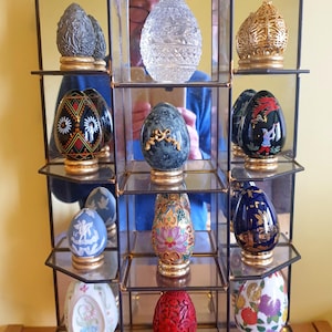 Puede incluir: Una vitrina de cristal llena de adornos decorativos con forma de huevo. Los huevos varían en color y diseño, incluyendo estilos tallados, pintados y enjoyados. Cada huevo descansa sobre una base dorada, creando una colección de objetos de colección ornamentados.