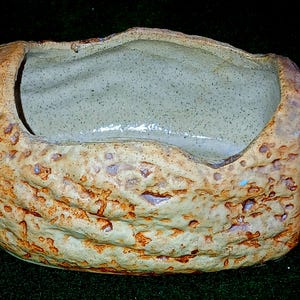 May include: A rectangular ceramic planter with a textured, rough exterior. The planter has a light brown and tan colour scheme with a light green interior. The edges are uneven, giving it a rustic appearance. Suitable for small plants or succulents.