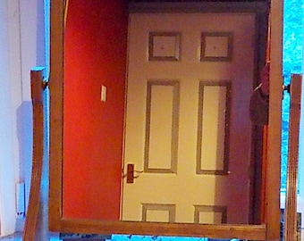May include: A wooden framed vanity mirror with a beveled edge and a shelf below. The mirror reflects a white door with a red wall behind it.