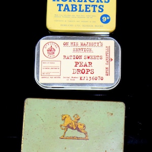 Puede incluir: Tres latas de metal vintage. La lata superior es amarilla con texto azul que dice "Horlicks Tablets 9d". La lata del medio es plateada con texto rojo y negro que dice "On His Majesty's Service. Ration Sweets Pear Drops KJ136070". La lata inferior es verde con una imagen dorada de una persona montando a caballo.